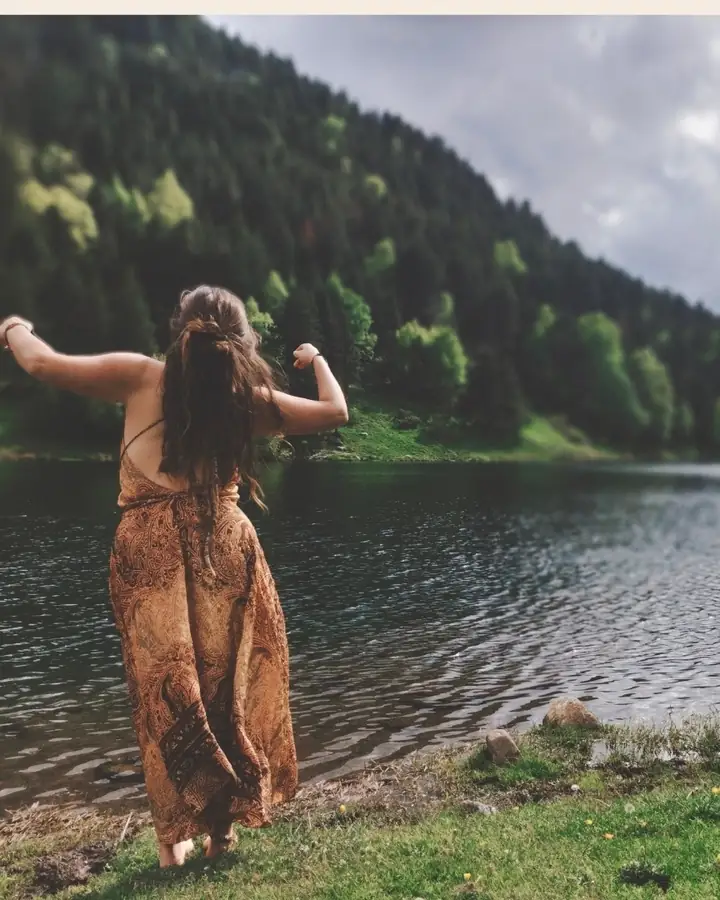 Une femme de dos, robe fluide et bras ouverts, danse face à un lac de montagne bordé de forêts