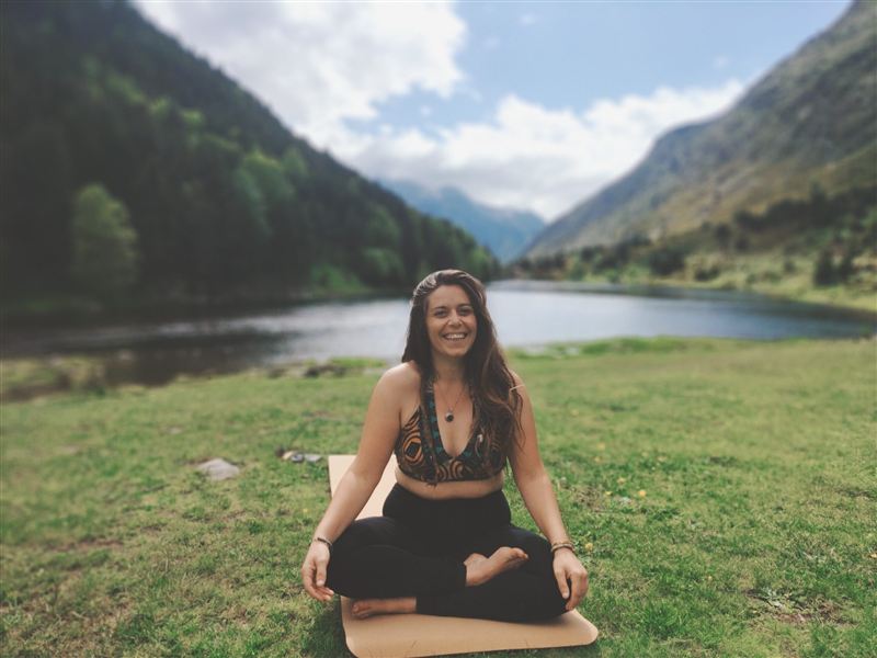 Pauline, souriante, assise en tailleur sur son tapis au bord d’un lac de montagne